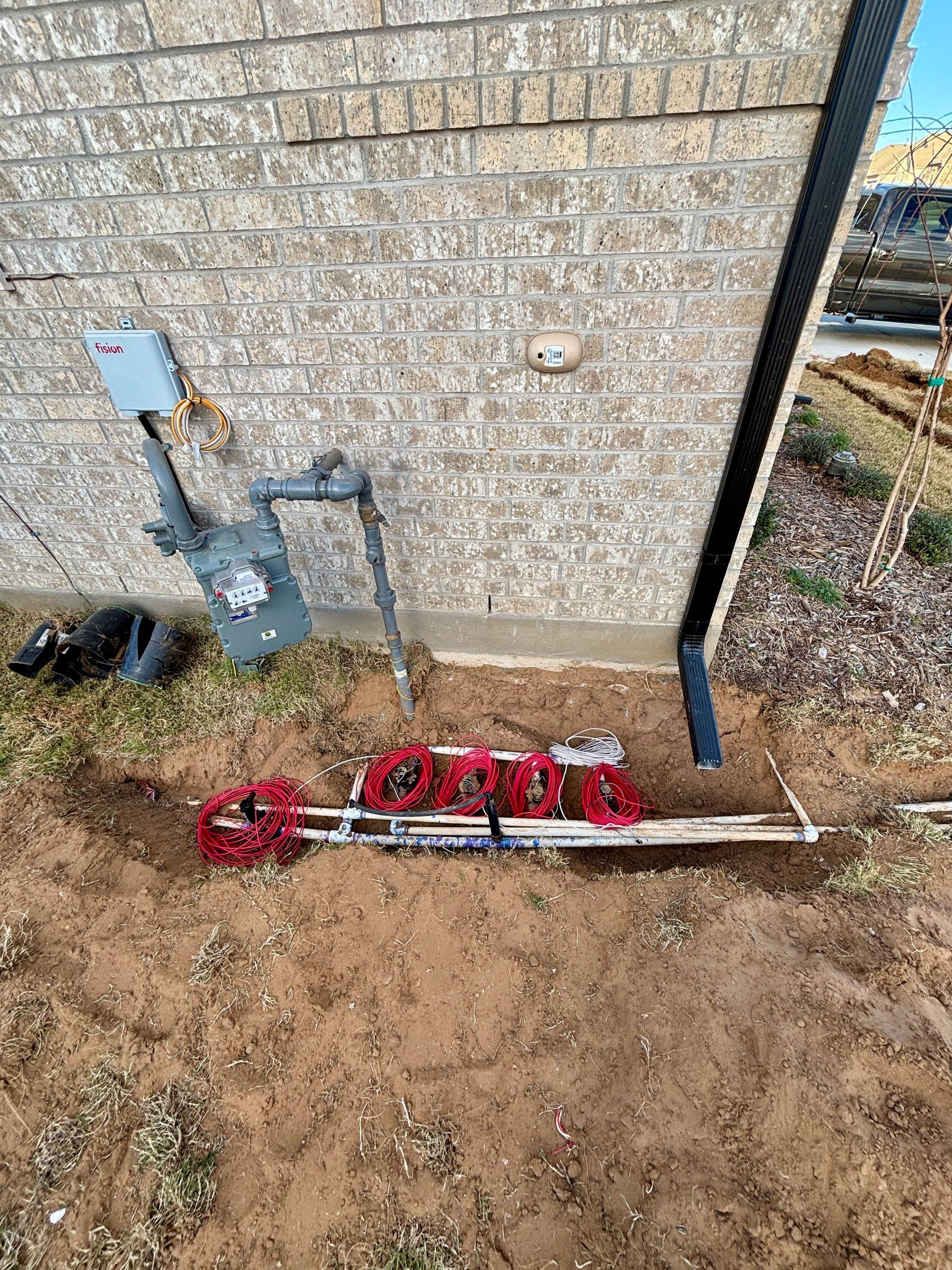Red utility valve and pipes beside a stone house wall on bare dirt ground.