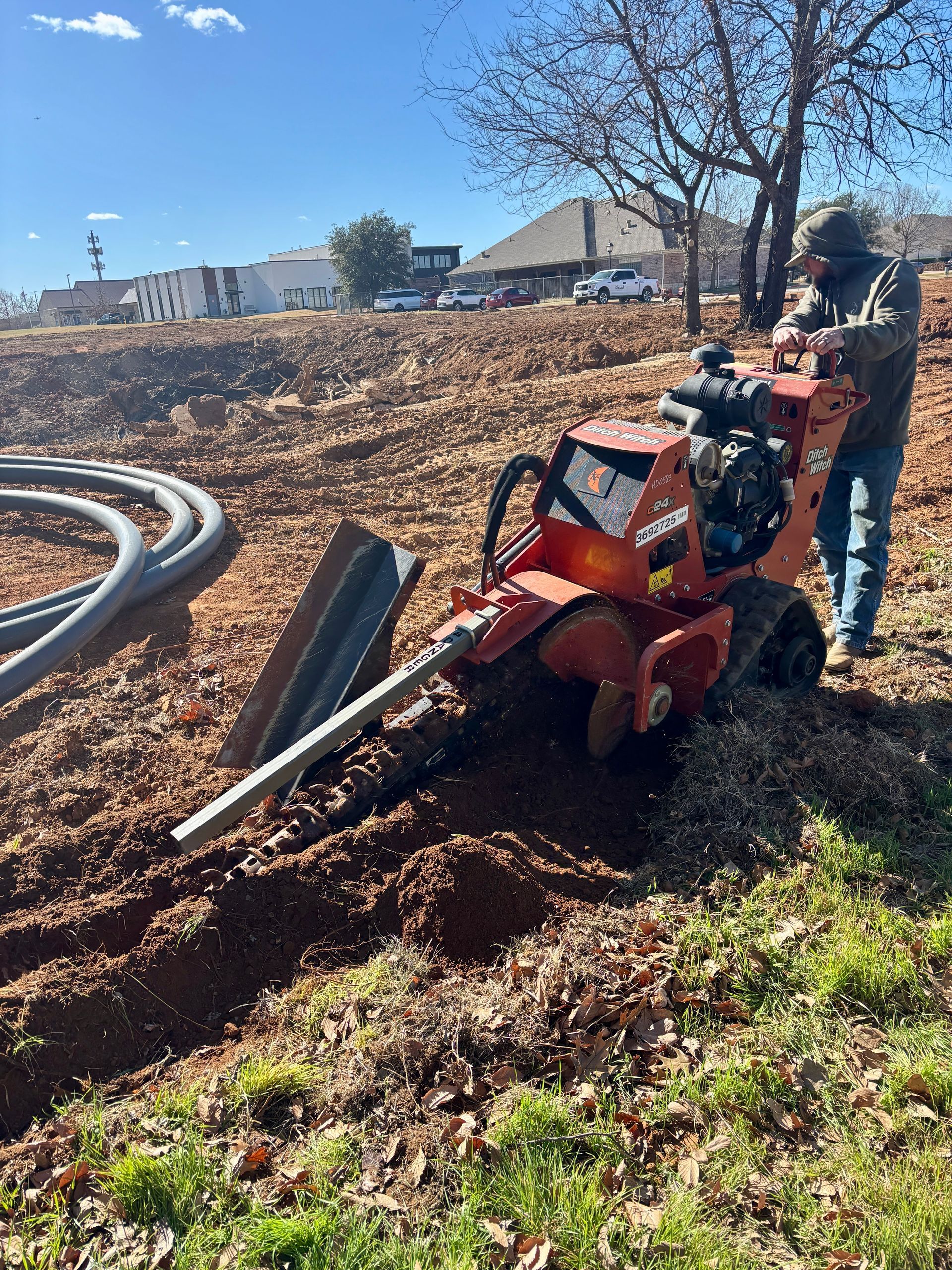 Person using a red mini excavator on a dirt lot beside a curved pipe, with a worker standing nearby.