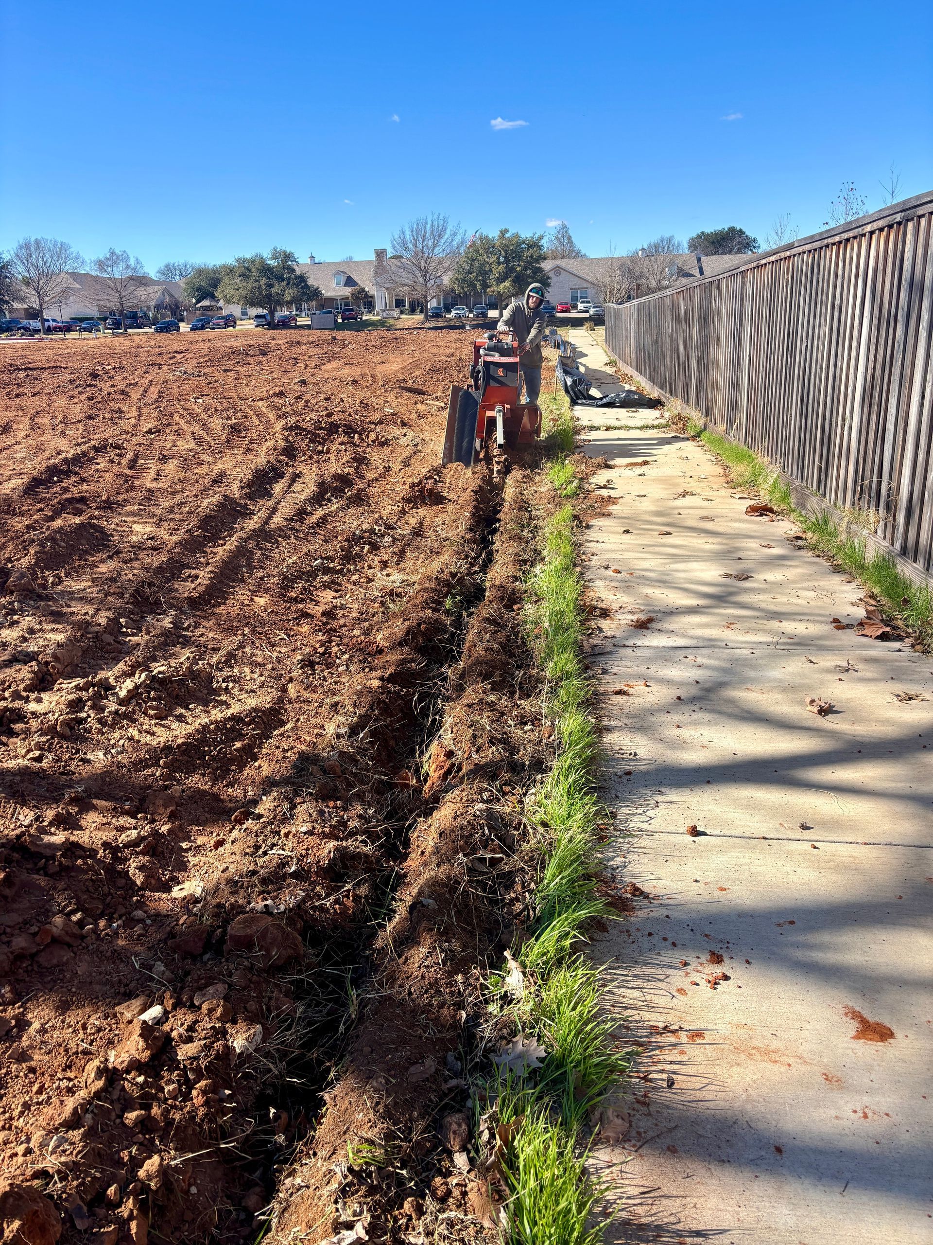 Sidewalk beside a tilled field and fence under a clear blue sky, with a person in orange in the distance