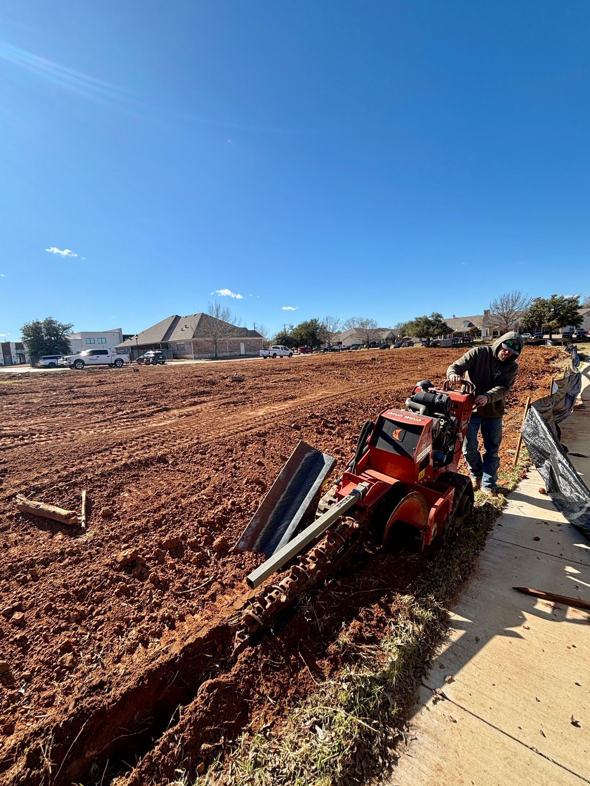 Red tractor stuck in a mud-filled ditch beside a sidewalk under a clear blue sky
