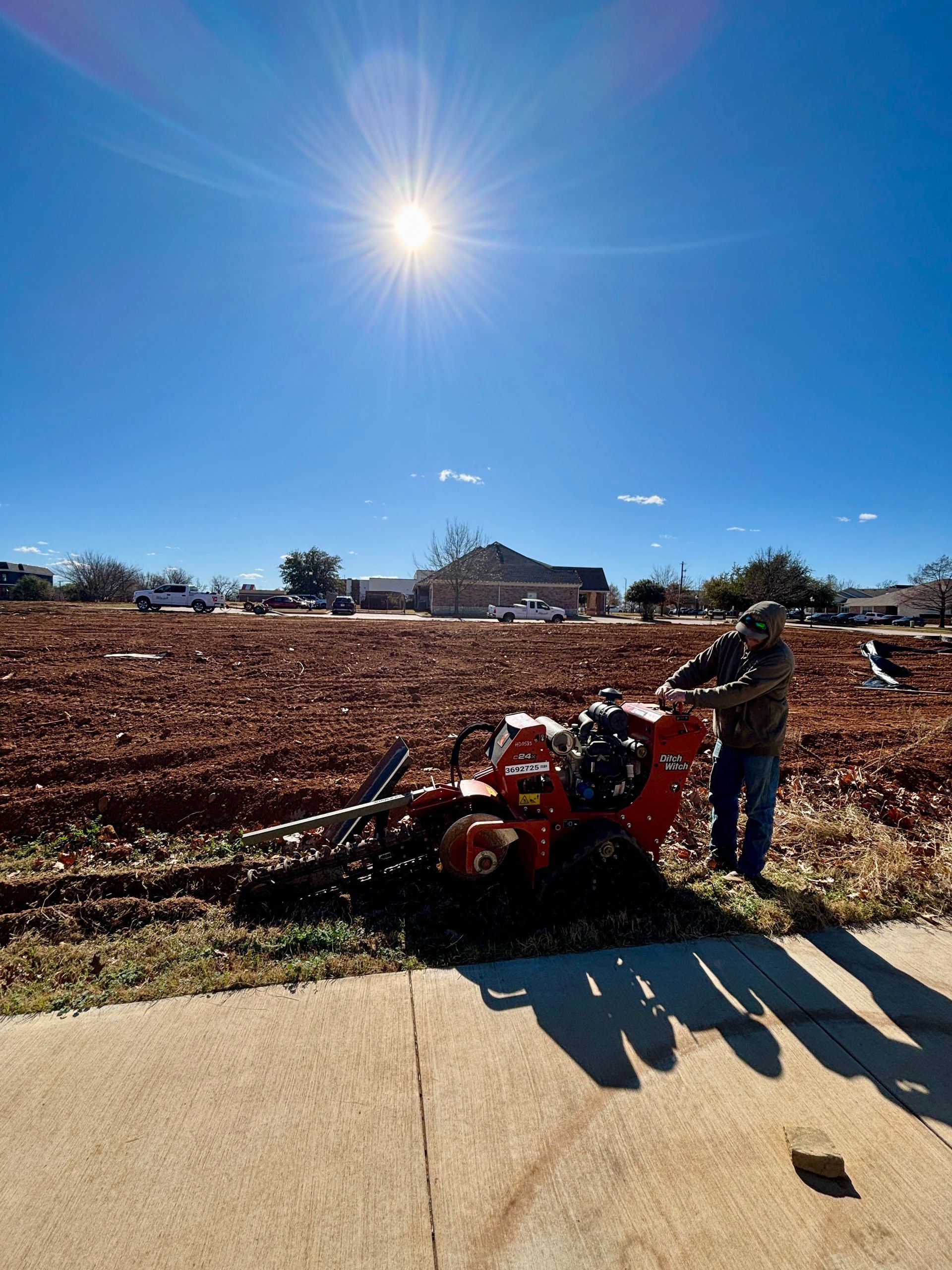 Person using a red tiller in a sunny field, with a bright blue sky and long shadows.