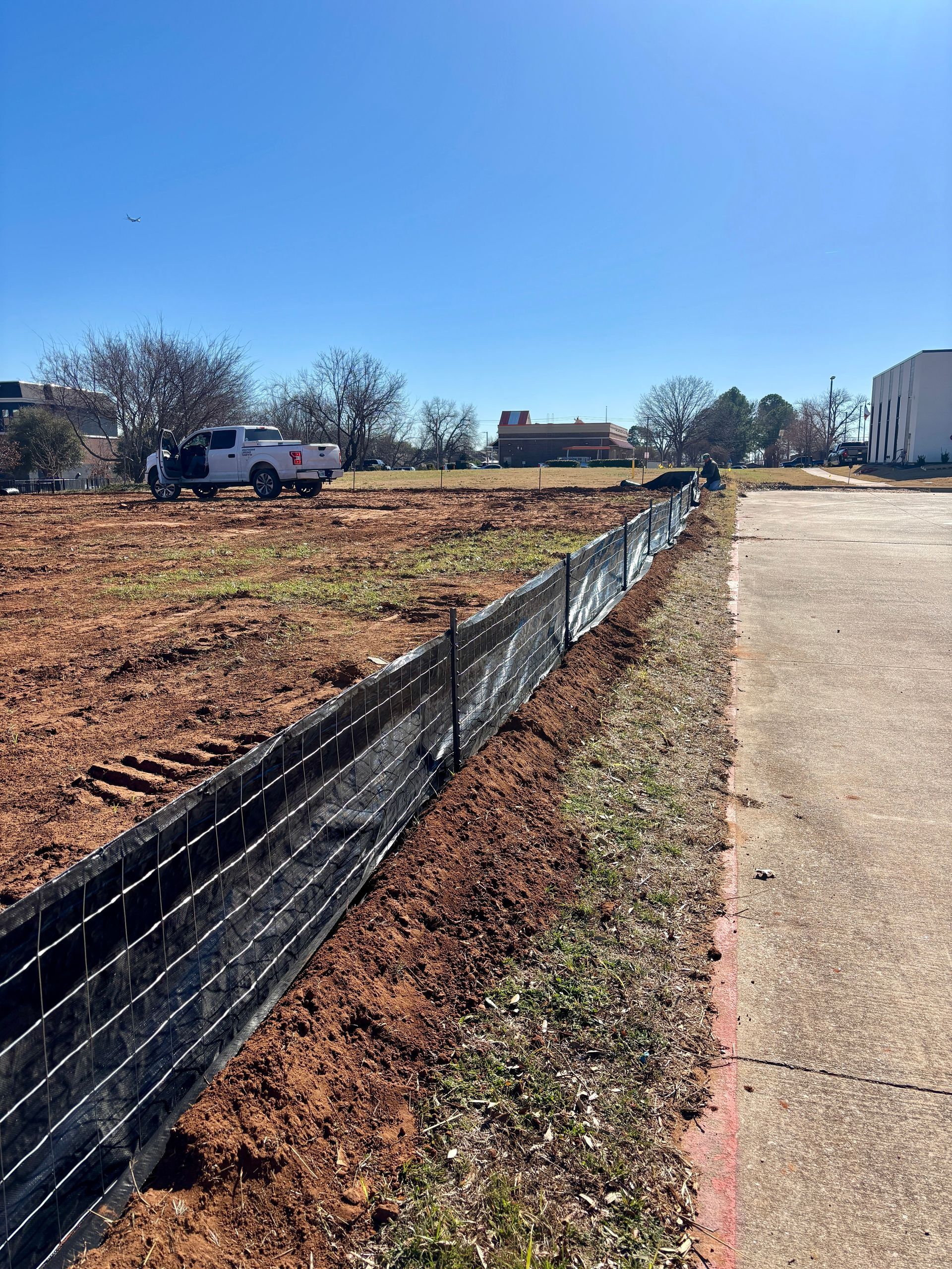 Empty lot beside a sidewalk, with a black fence, pickup truck, and buildings under a clear blue sky.