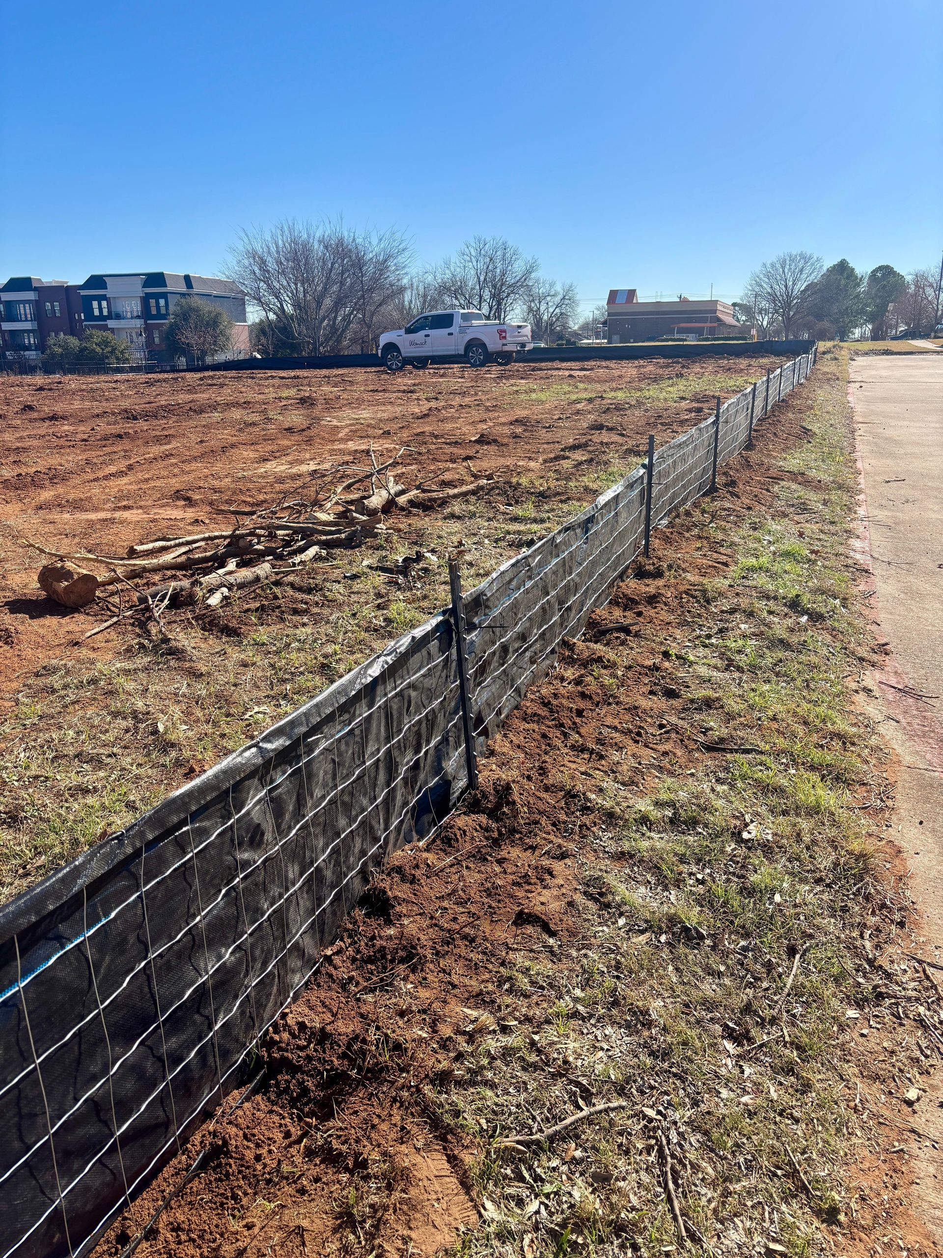 Bare dirt lot beside a gravel path, with a black silt fence and a white pickup in the distance.