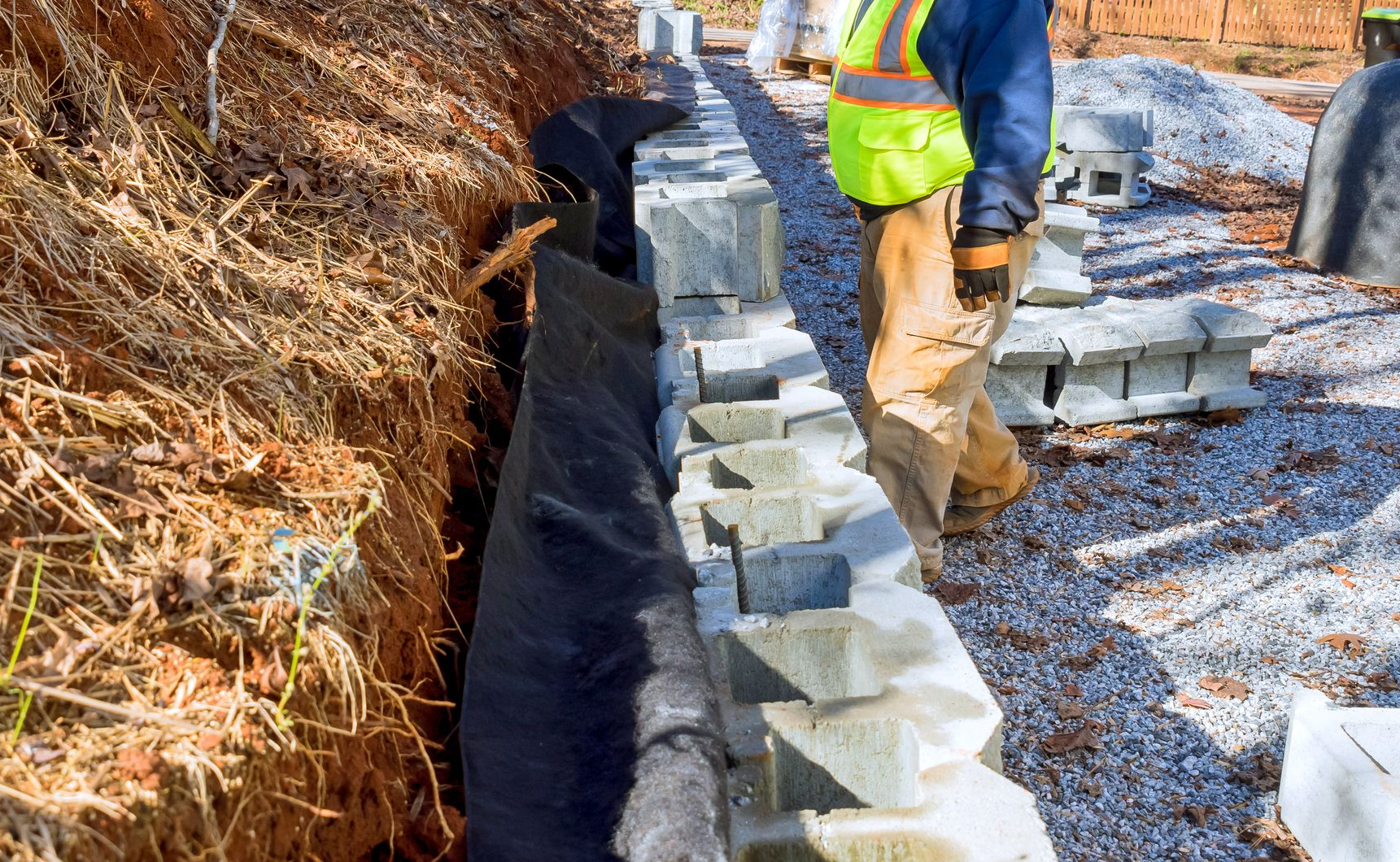 Construction worker stacking concrete blocks beside an excavated trench on a gravel site.