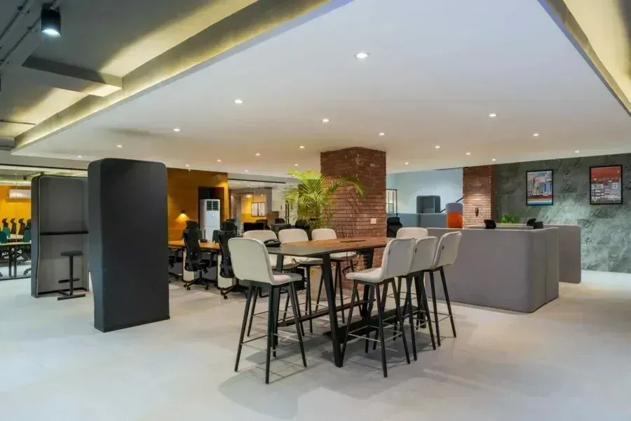 Modern office lounge with high tables, stools, gray seating, and warm lighting in an open workspace