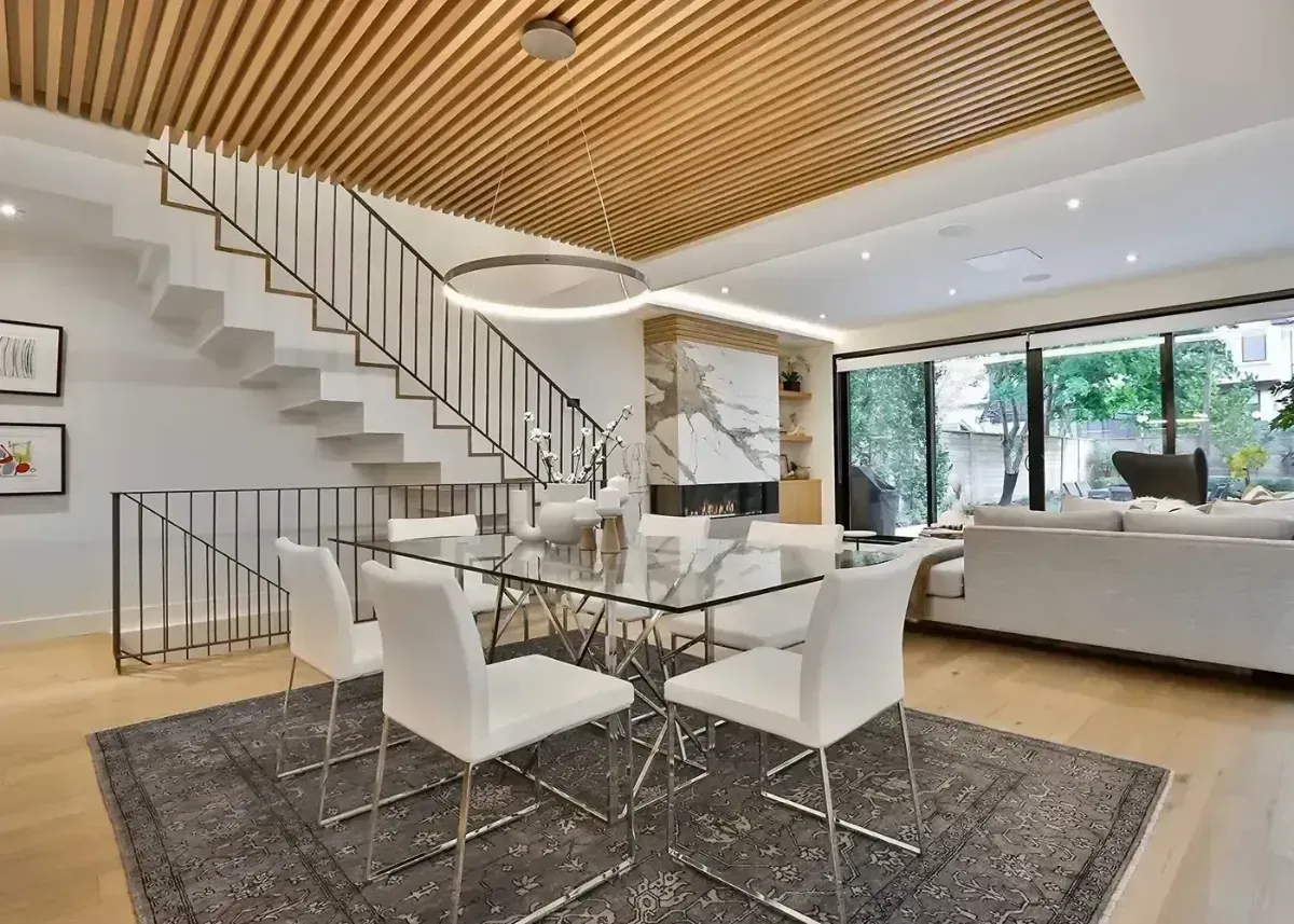 Modern open-plan living room with staircase, dining table, and large windows, featuring wood ceiling and white furniture