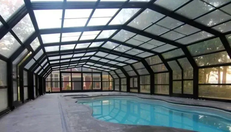 Indoor swimming pool under a glass-enclosed roof with a covered patio and lounge area