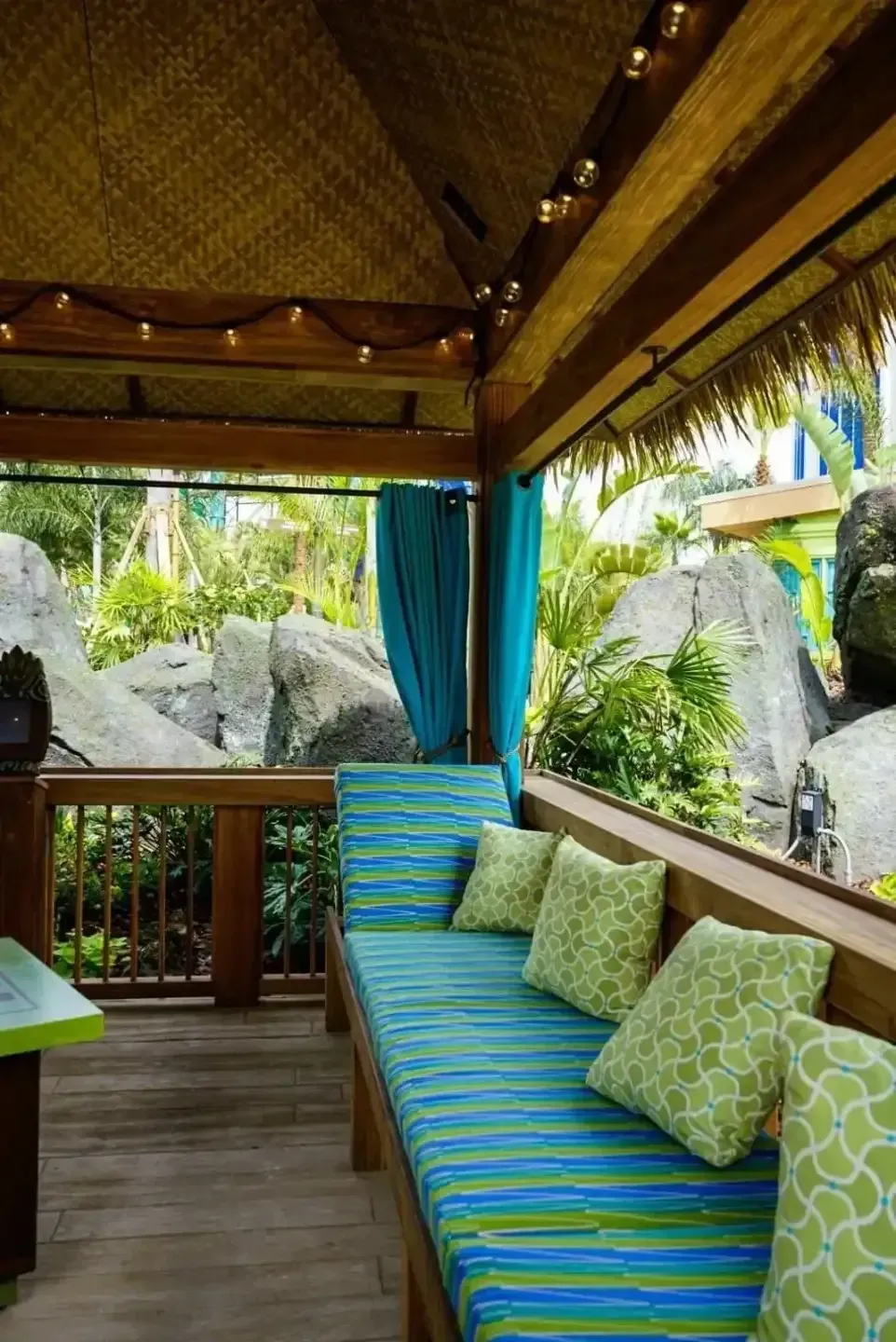 Shaded tropical patio with blue-striped daybed, green pillows, and wooden railing overlooking rocks and palm trees