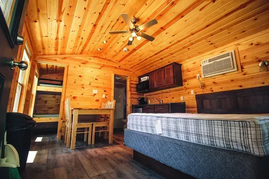 Cozy log cabin bedroom with wood-paneled walls, bed, desk, chair, ceiling fan, and window AC unit