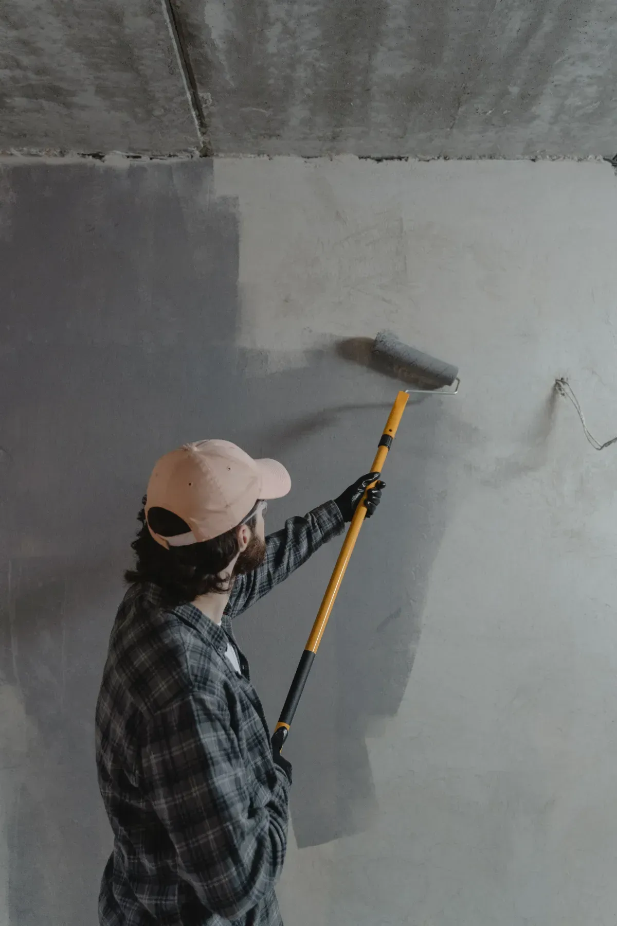 Person painting a gray wall with a roller on a telescoping pole in an indoor room.
