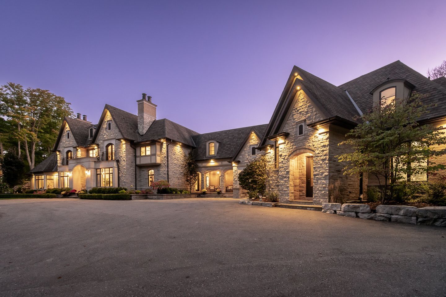 Stone mansion at dusk with warm-lit windows, peaked roofs, and a wide gravel driveway