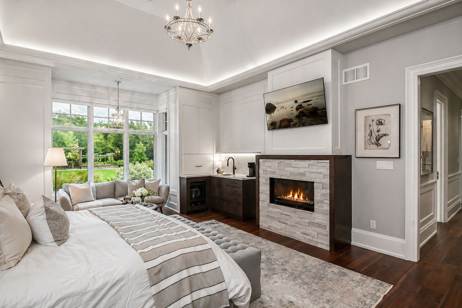 Bright bedroom with fireplace, gray walls, striped bedding, chandelier, and large window overlooking greenery