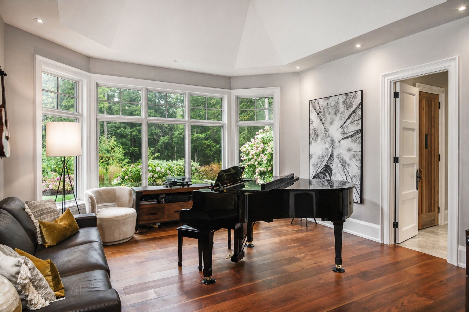 Bright music room with a grand piano, bay windows, hardwood floors, and a black leather sofa