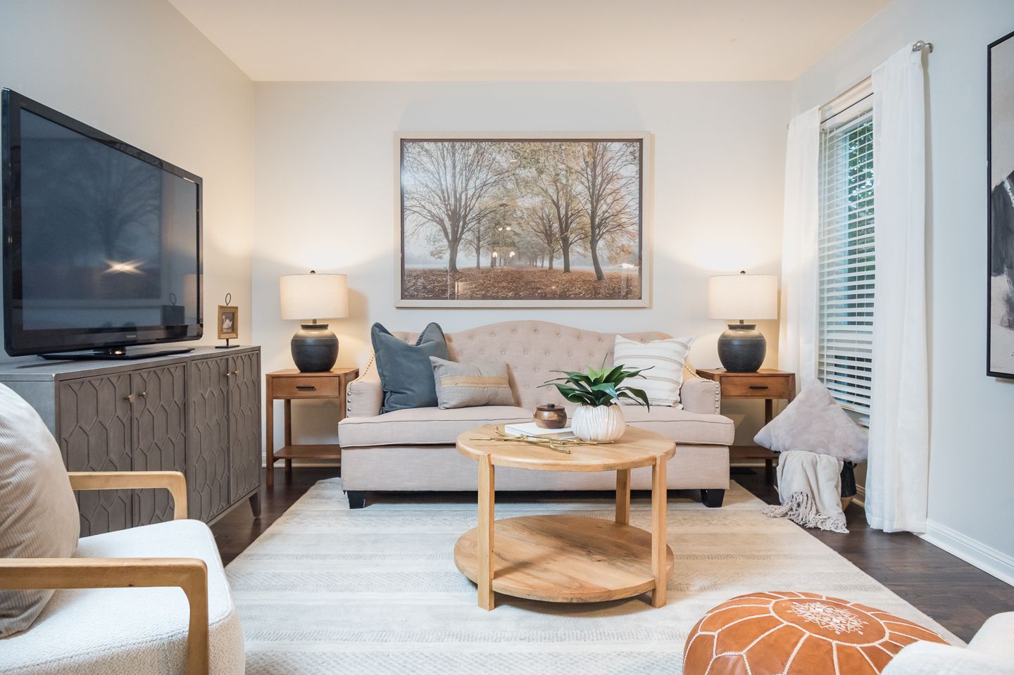 Bright living room with beige sofa, TV, coffee table, lamps, and large landscape wall art