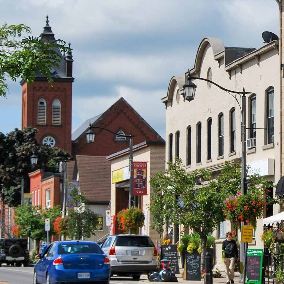 Downtown street with cars, storefronts, trees, and a red-brick church tower on a sunny day