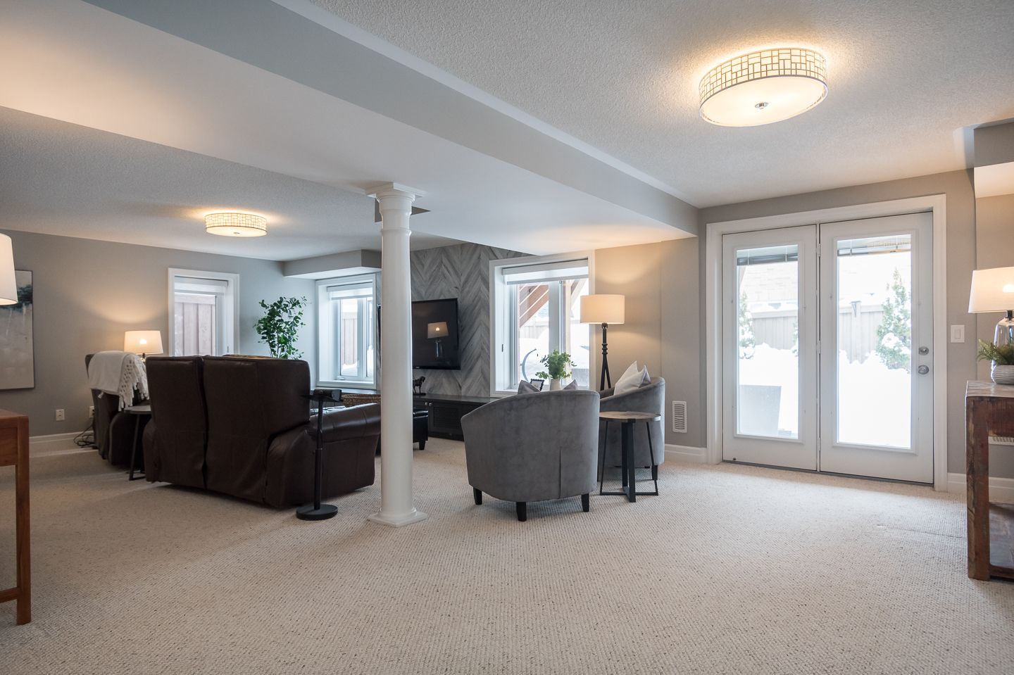 Spacious carpeted living room with gray and brown chairs, white columns, and French doors to a bright patio