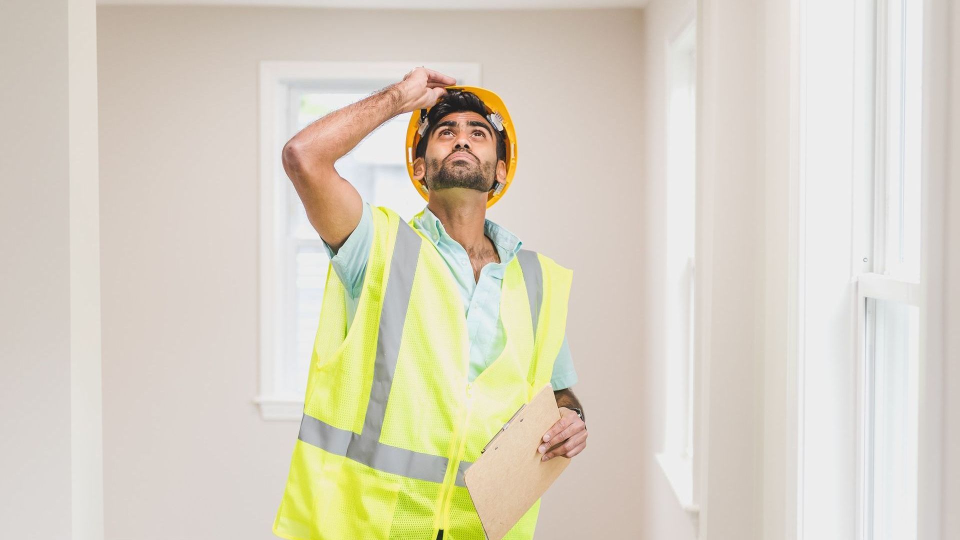 A man wearing a hard hat and safety vest is holding a clipboard and looking up.