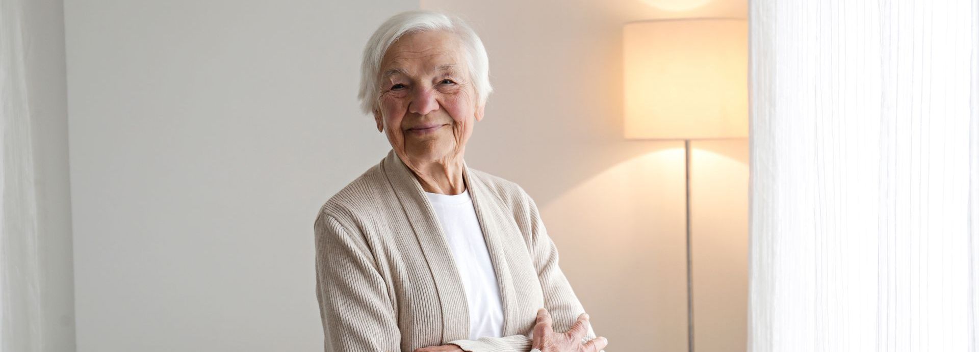 An elderly woman is standing in a living room with her arms crossed.