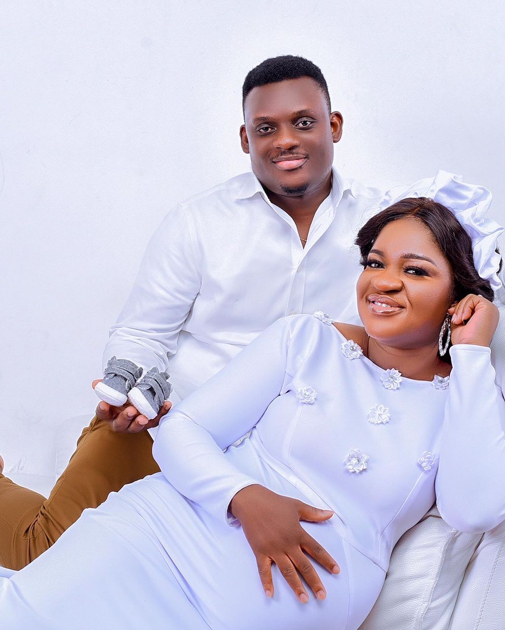 A man and a pregnant woman are posing for a picture while the woman is holding baby shoes.