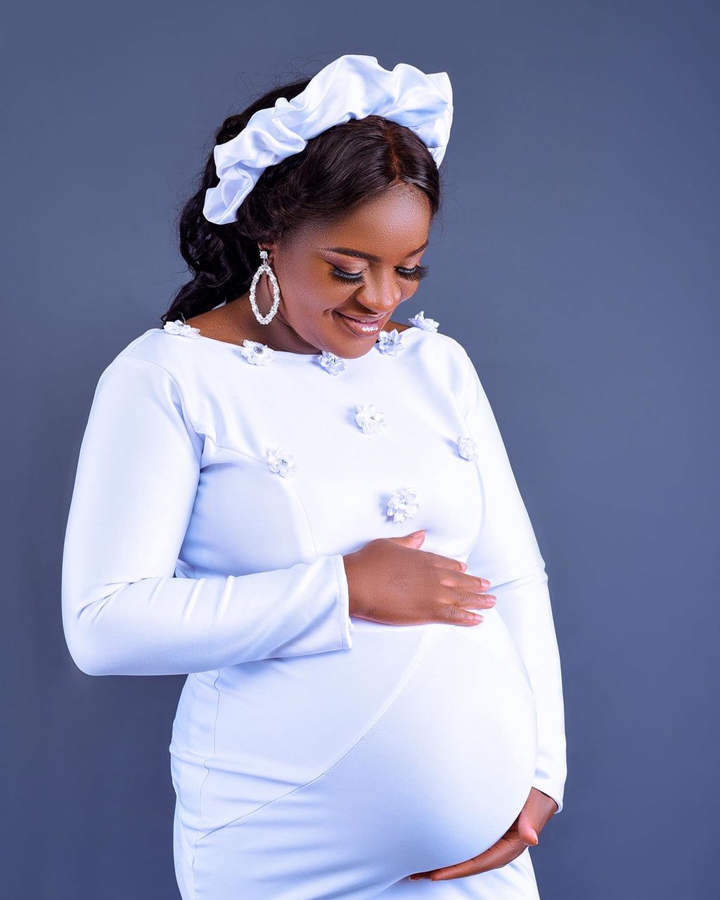 A pregnant woman in a white dress is holding her belly.