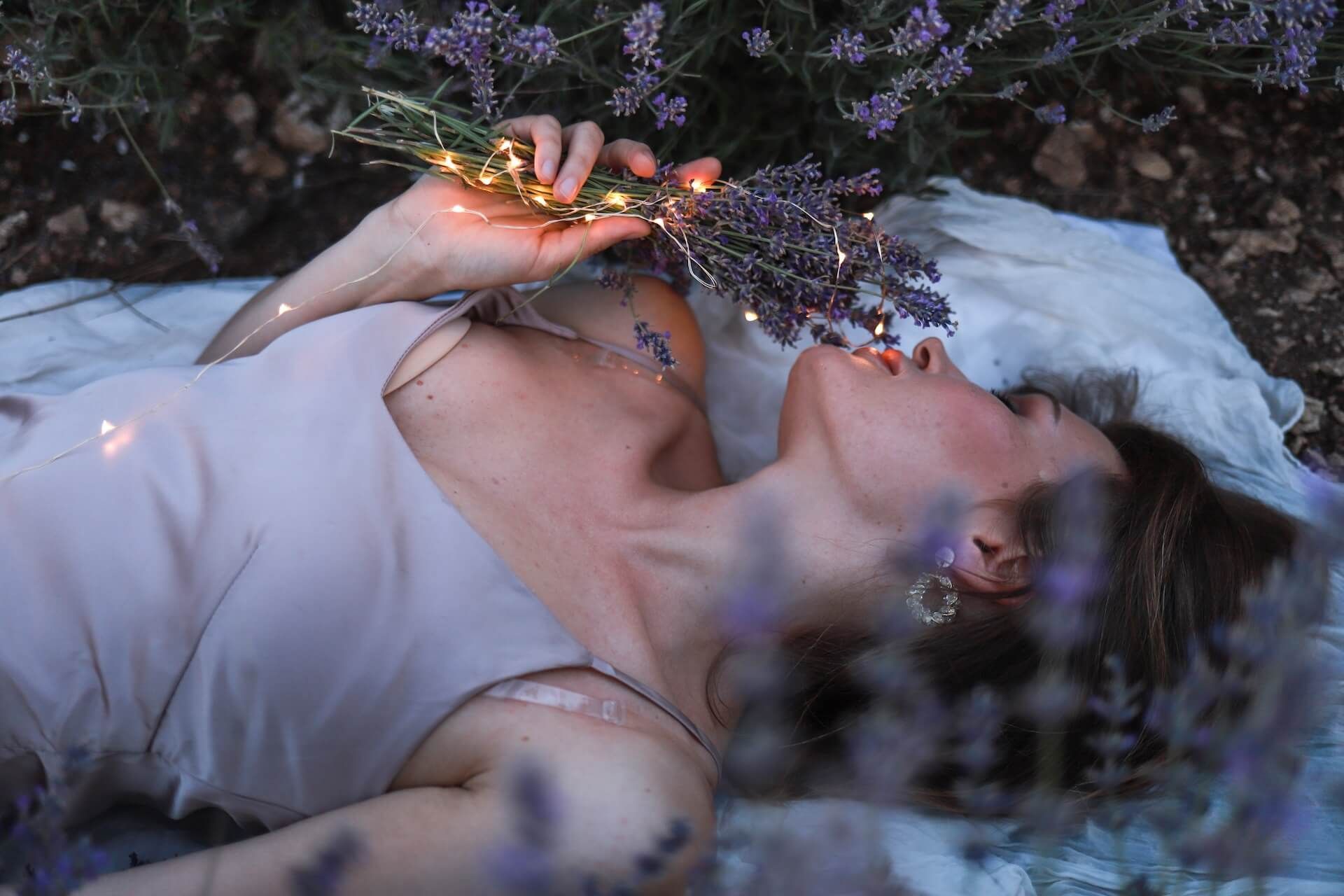 Woman lying in lavender field, holding lit flowers, eyes closed.