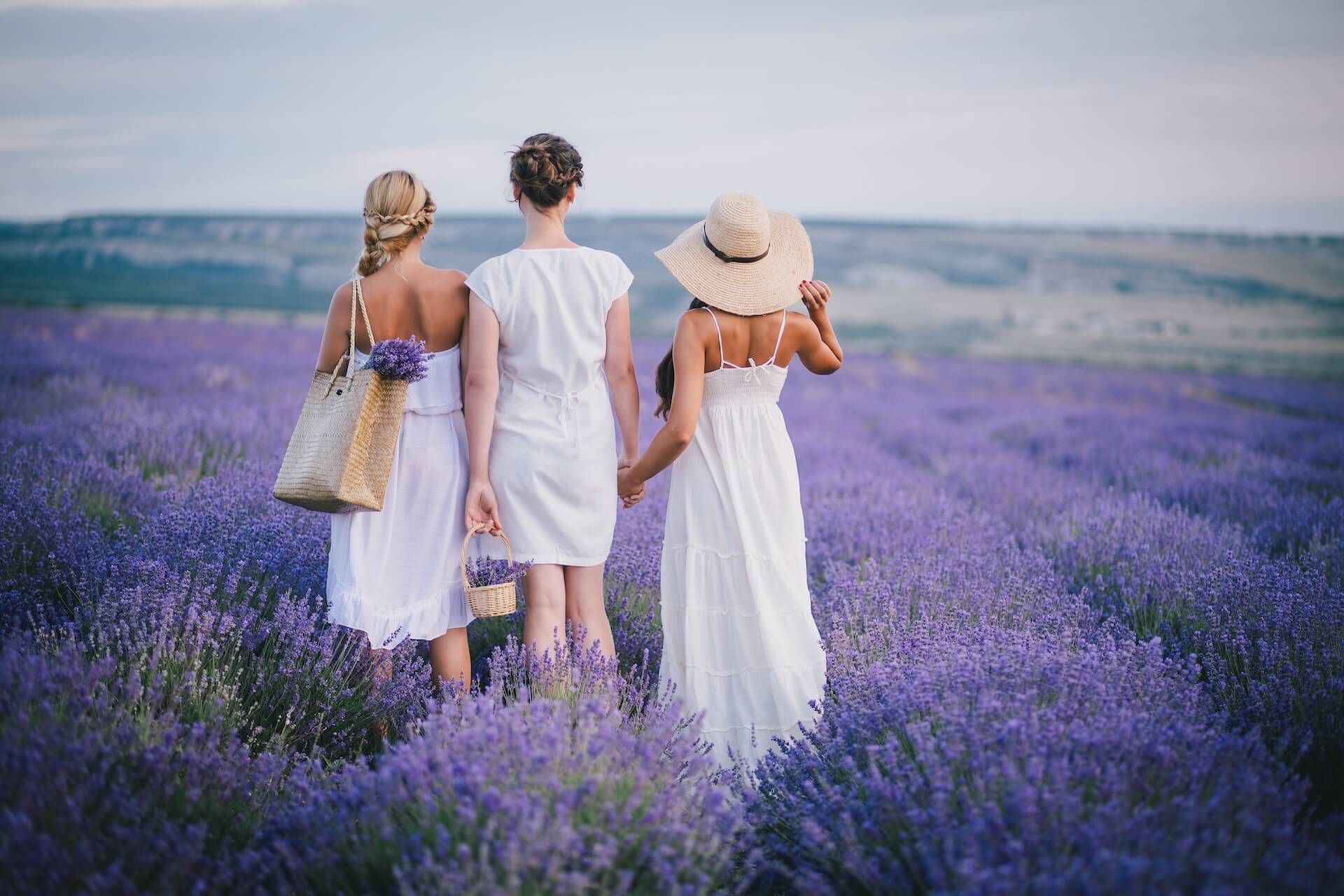 Three women in white dresses holding hands in a lavender field; one wears a straw hat and carries a tote.