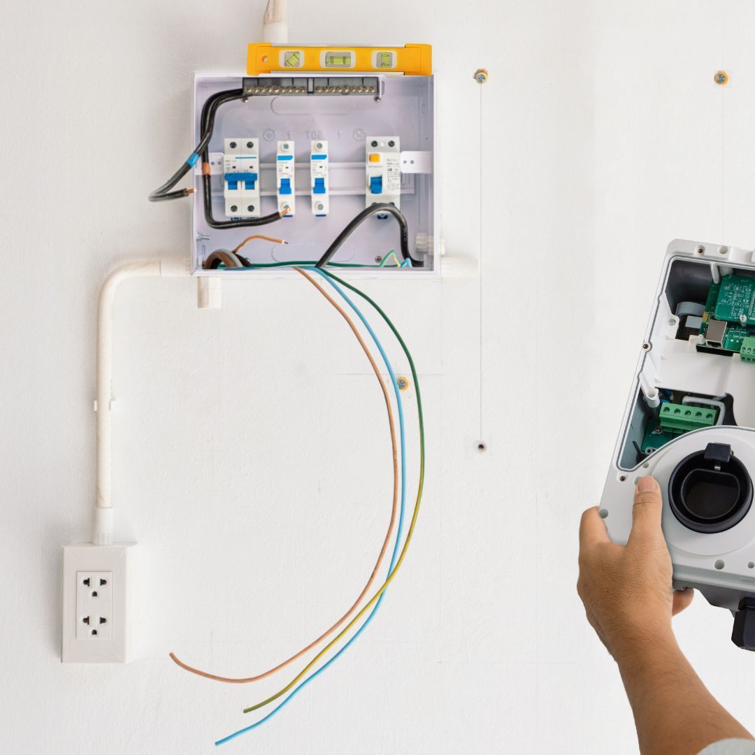 Electrician wiring a wall breaker panel, with exposed colored cables and a hand holding an electrical device.