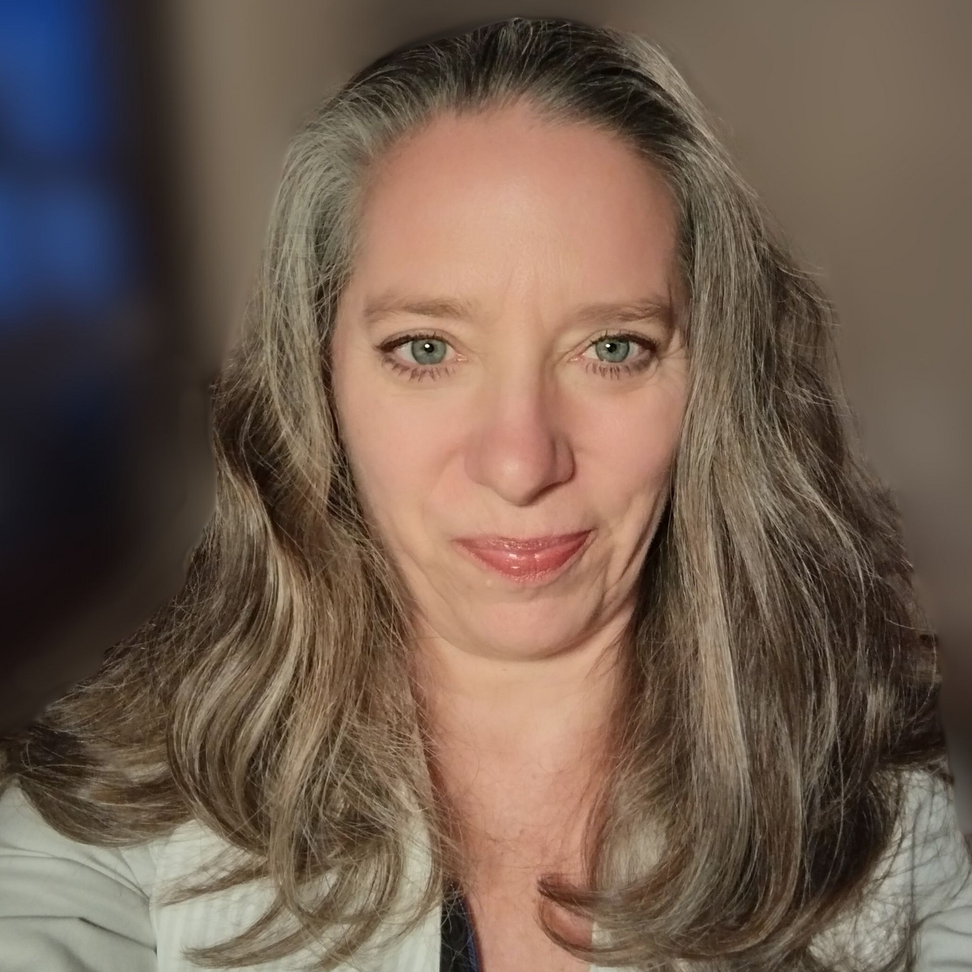 Woman with long, wavy gray hair, wearing a white blazer, looking at the camera with a neutral expression.