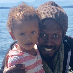 Man with a child, both smiling, by the water; man wearing a hat.