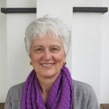 Woman with short gray hair, wearing a purple scarf and gray shirt, smiling.