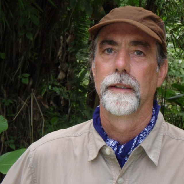 Man with gray beard, wearing a brown cap and tan shirt, stands in a lush green setting.