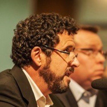 Man with curly hair and beard, wearing glasses, speaking into a microphone. Another man visible behind him.