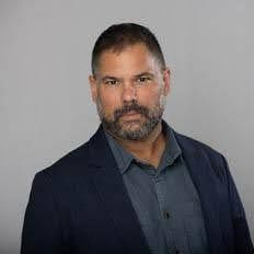 Man with short hair and beard wearing a navy blazer and blue button-down shirt. Gray background.