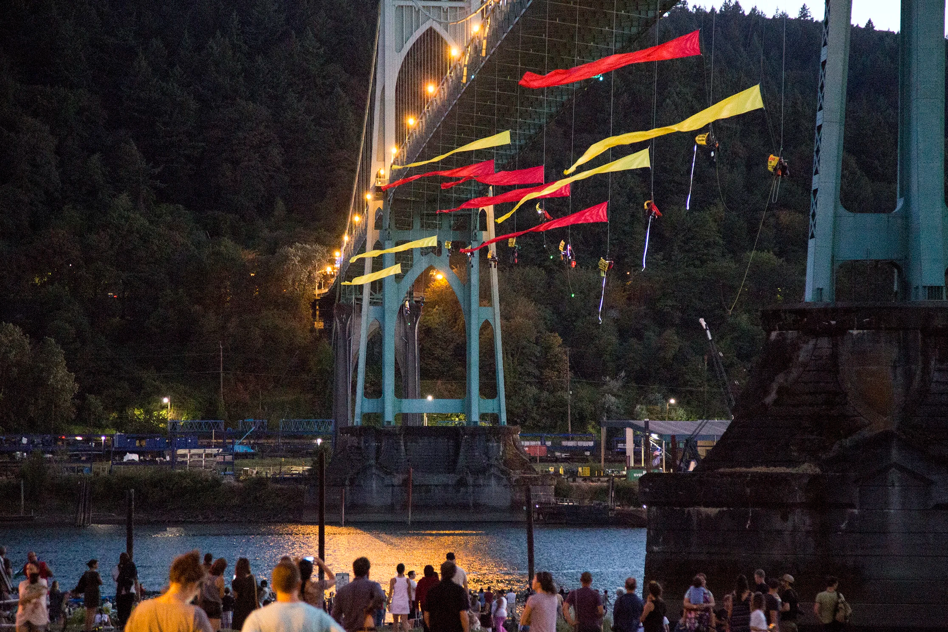 St. Johns Bridge in Portland, OR, decorated with colorful flags, at dusk, with people gathered below.