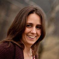 Woman with brown hair smiling, wearing a dark sweater outdoors with a blurred background.
