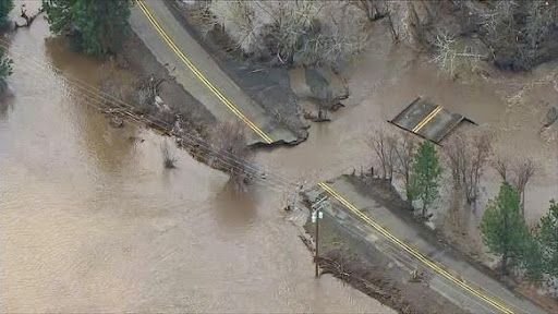 Flooded road, sections collapsed. Brown water surrounds broken pavement and trees.