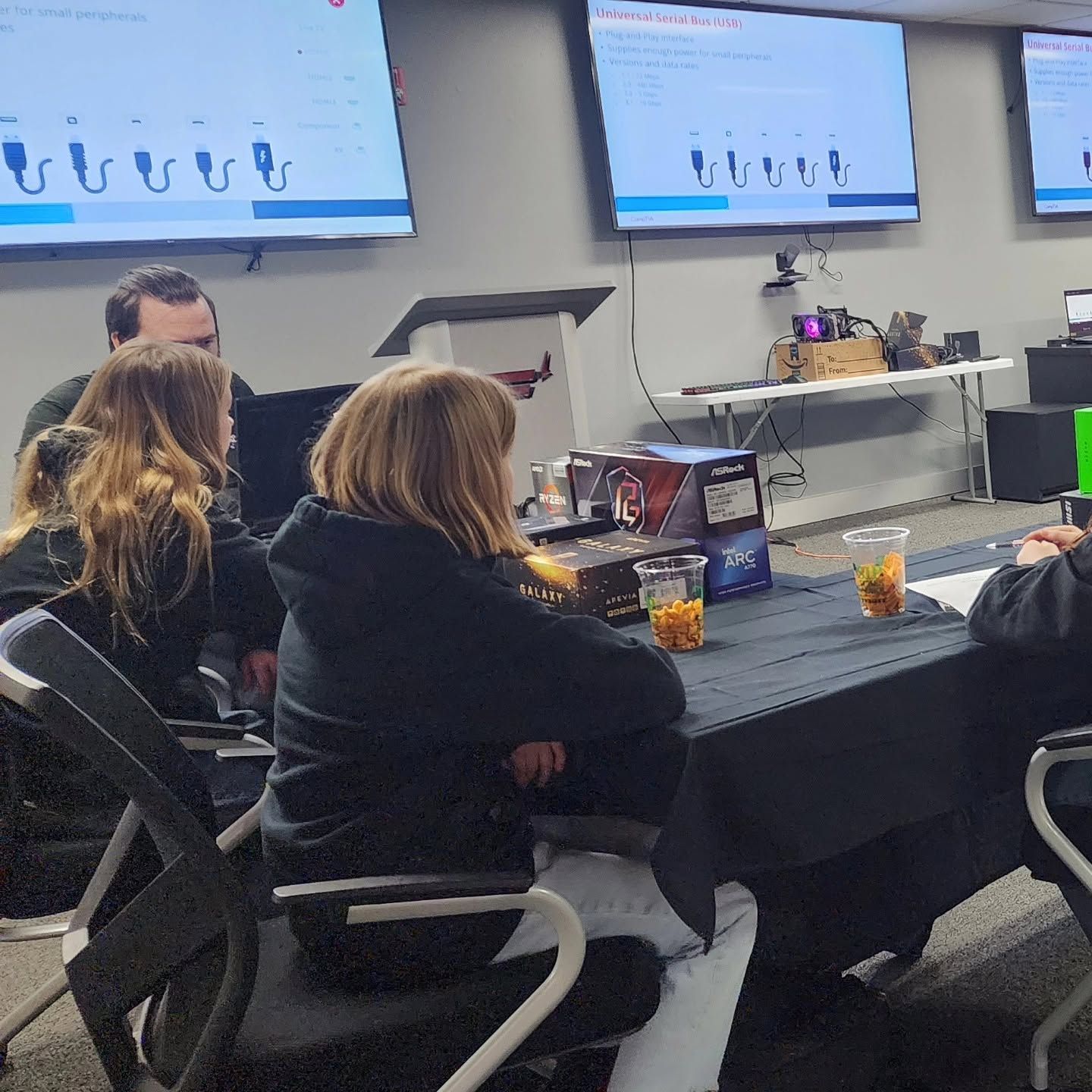 People seated at a conference table in a meeting room watching a presentation on wall screens.
