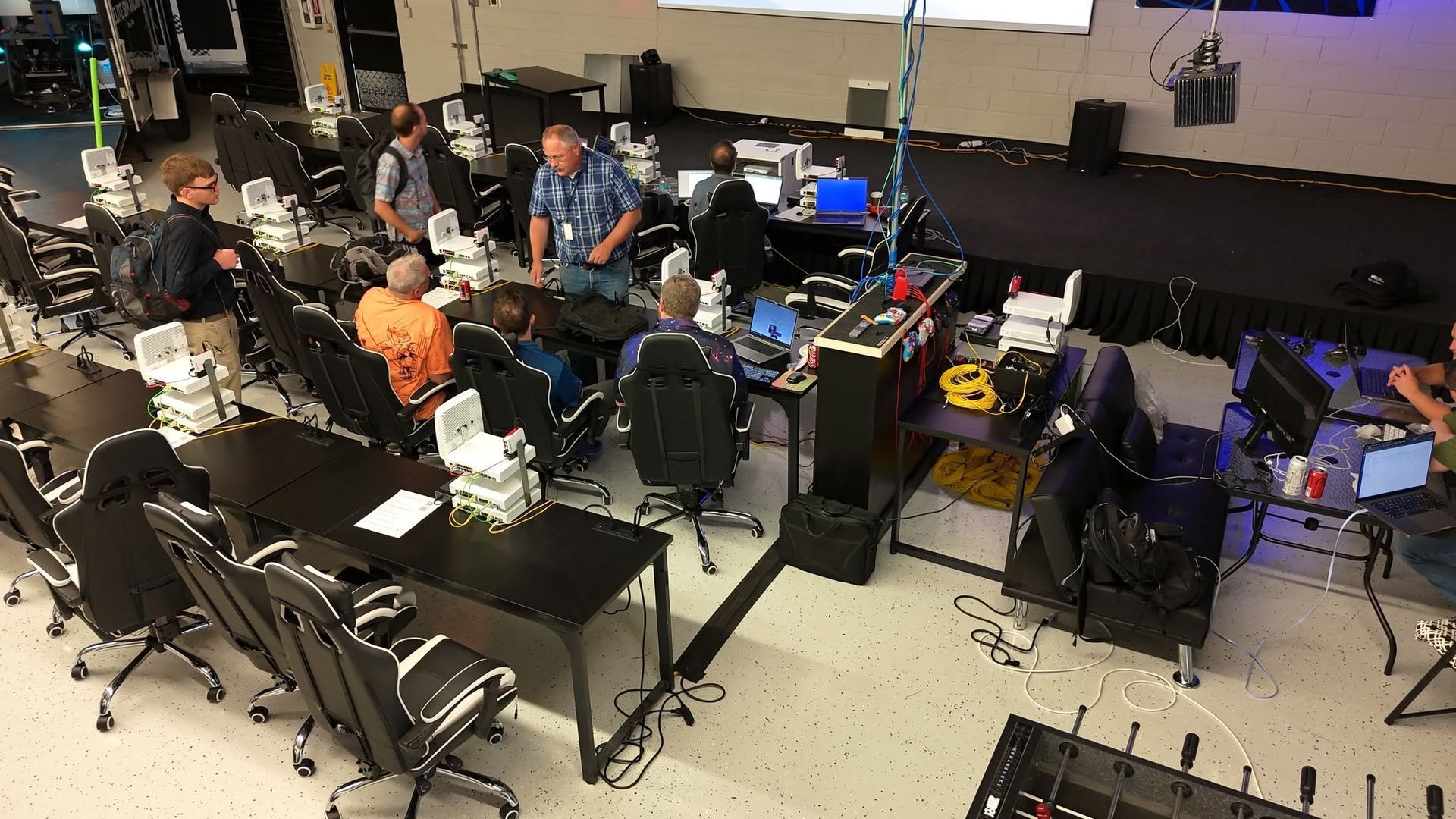 People seated around computer stations in a busy classroom or lab, with cables, monitors, and equipment scattered around.