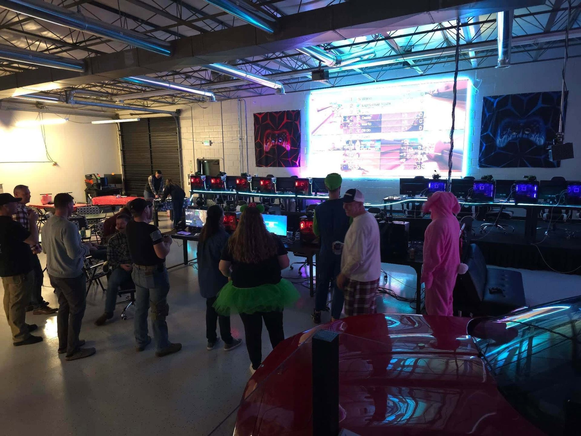People gathered in a neon-lit gaming event space with screens, computers, and a large projection display.