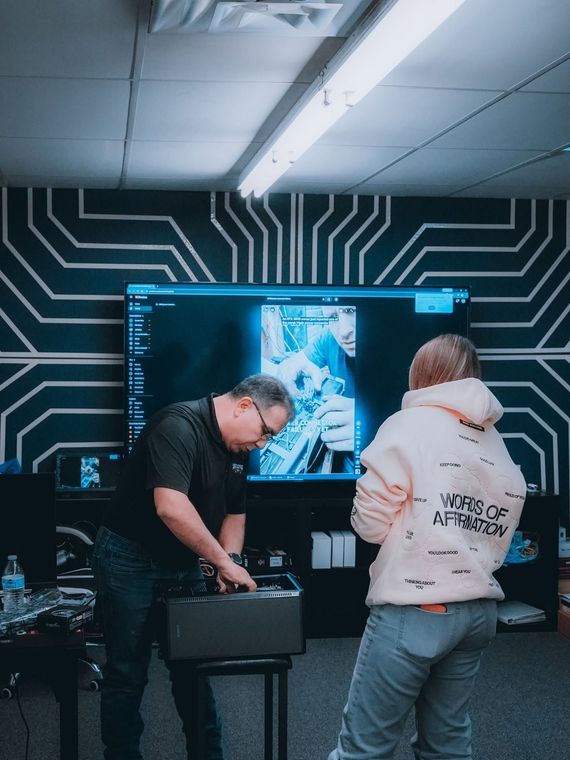 Two people collaborate in a tech lab beside a large monitor and circuitry-patterned wall