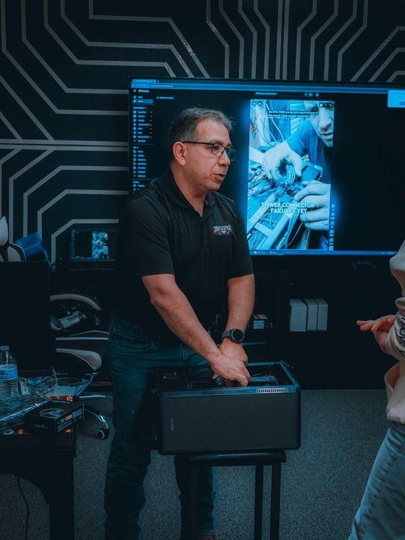 Man in black polo beside a server case in a tech room with monitors and circuit-like wall graphics