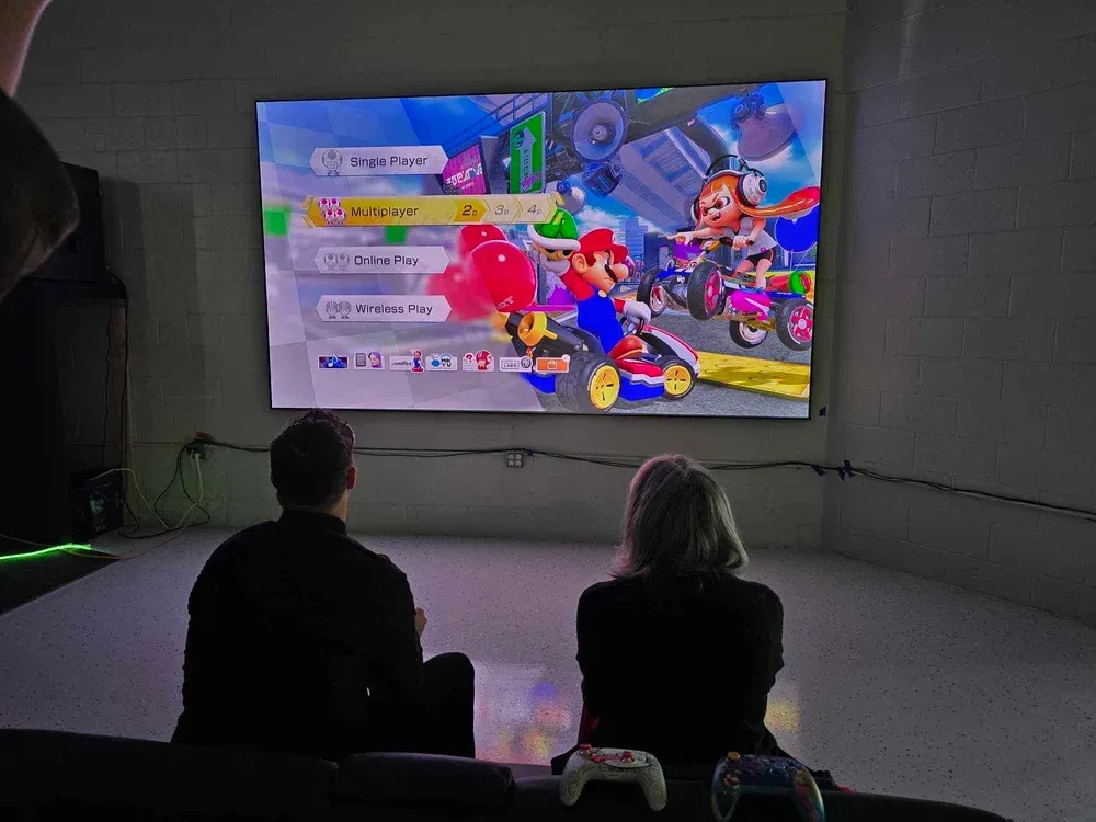 Two people sit facing a large screen showing a colorful Mario Kart race in a dark room.