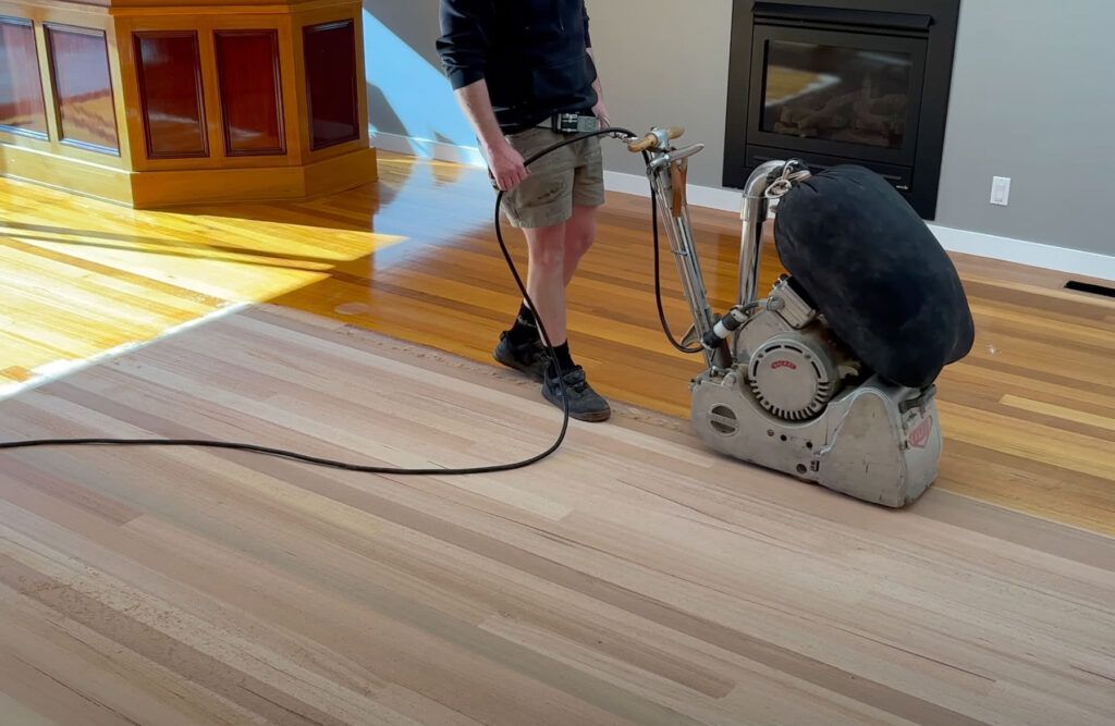 Man using a floor sander on a hardwood floor in a room with a fireplace and sunlight.