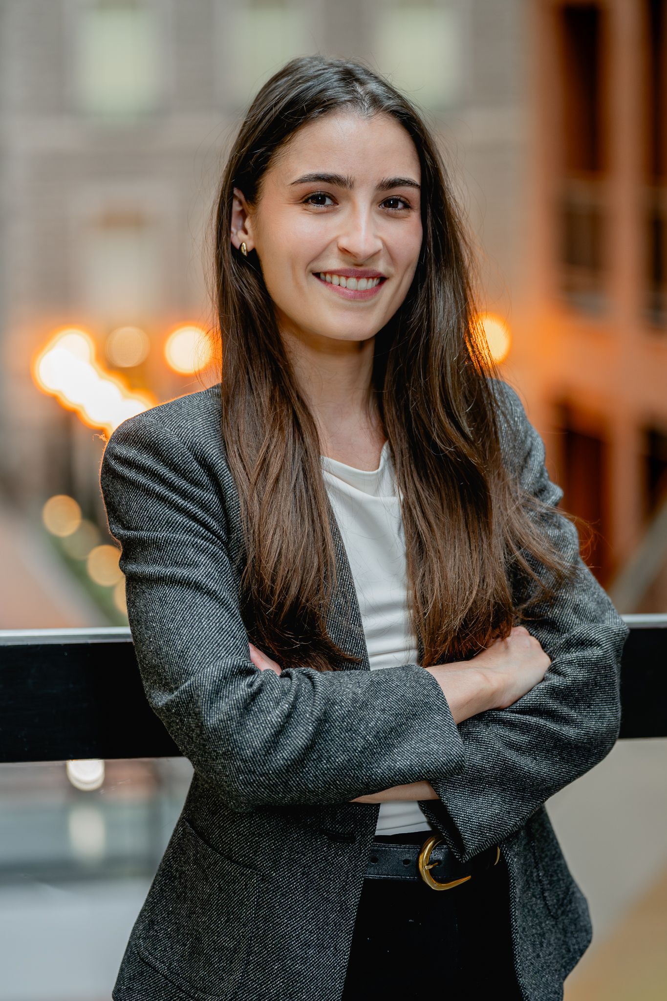 Femme souriante en blazer gris, les bras croisés, dans un décor urbain intérieur