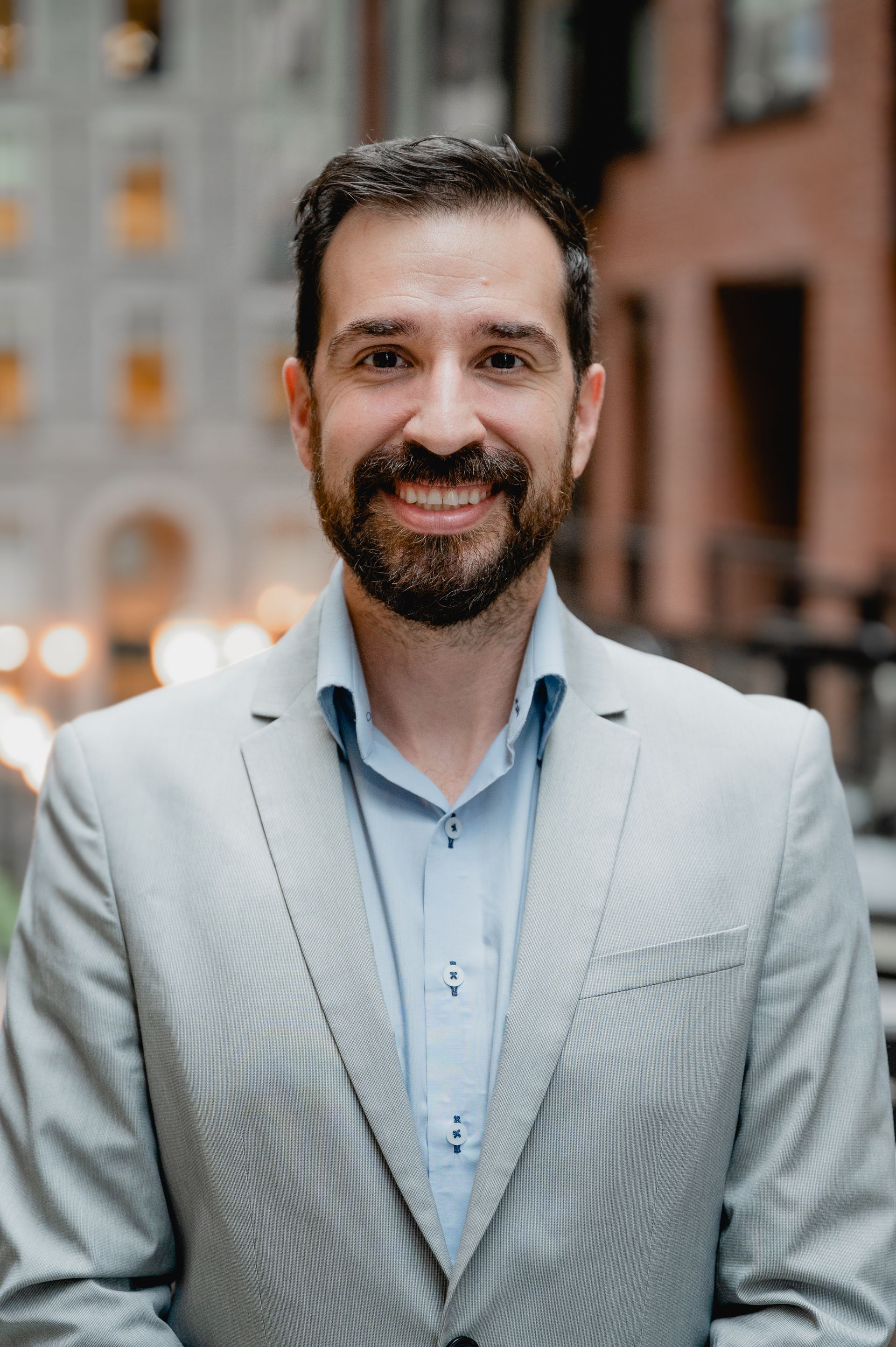 Un homme en blazer gris clair et chemise bleue sourit dans une rue de la ville.