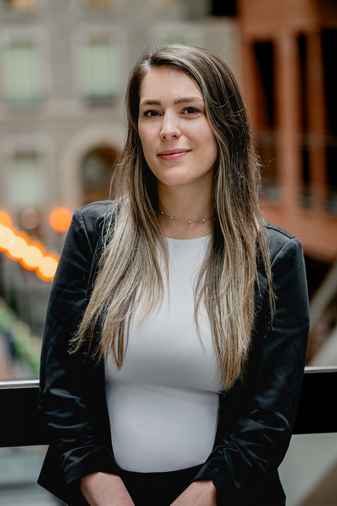 Femme en blazer noir et haut blanc, debout à l'extérieur, avec les lumières floues de la ville en arrière-plan.