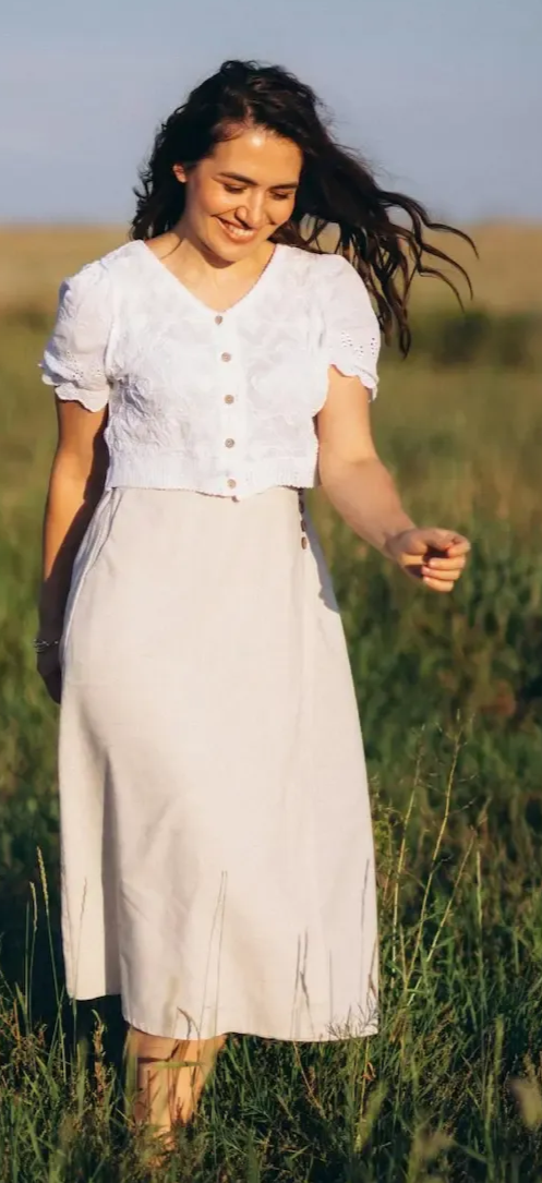 A person smiling while walking through a sunny field in a white top and a long, light-colored skirt.