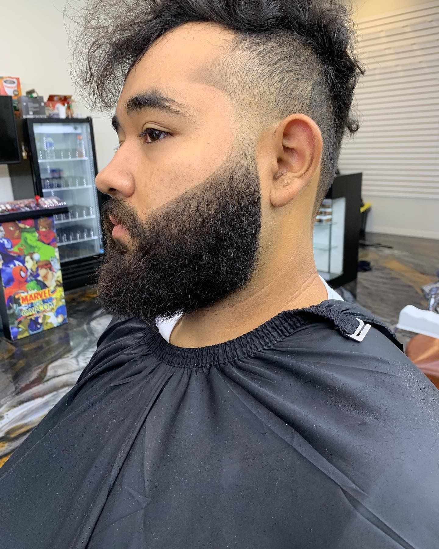 A man with a neatly faded haircut and a groomed, shaped beard sitting in a barber’s cape inside a salon.