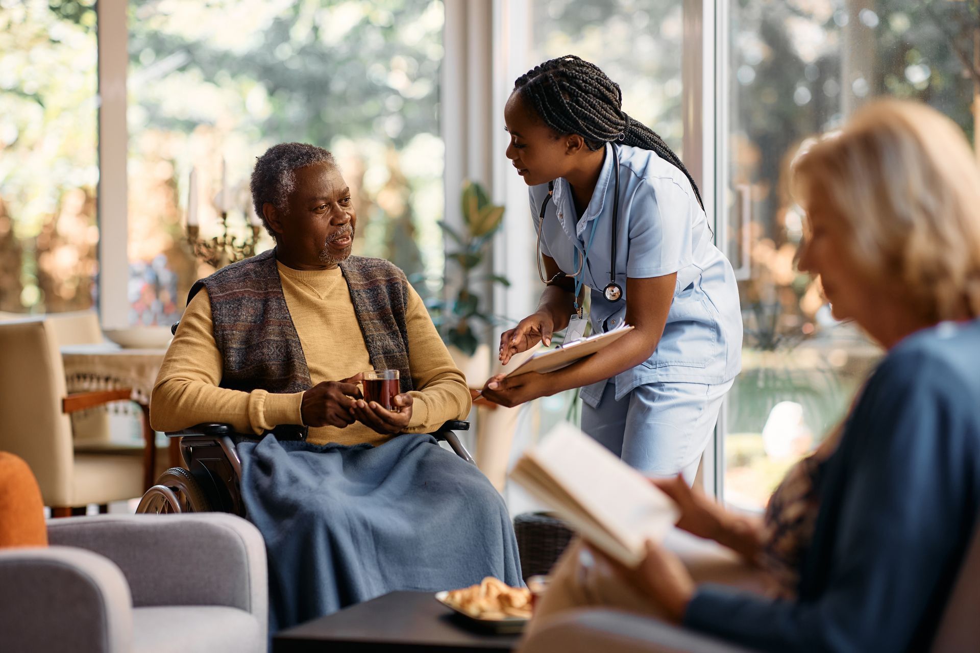 Nurse talks to elderly woman in wheelchair, another woman reads nearby. Bright, sunny living room setting.