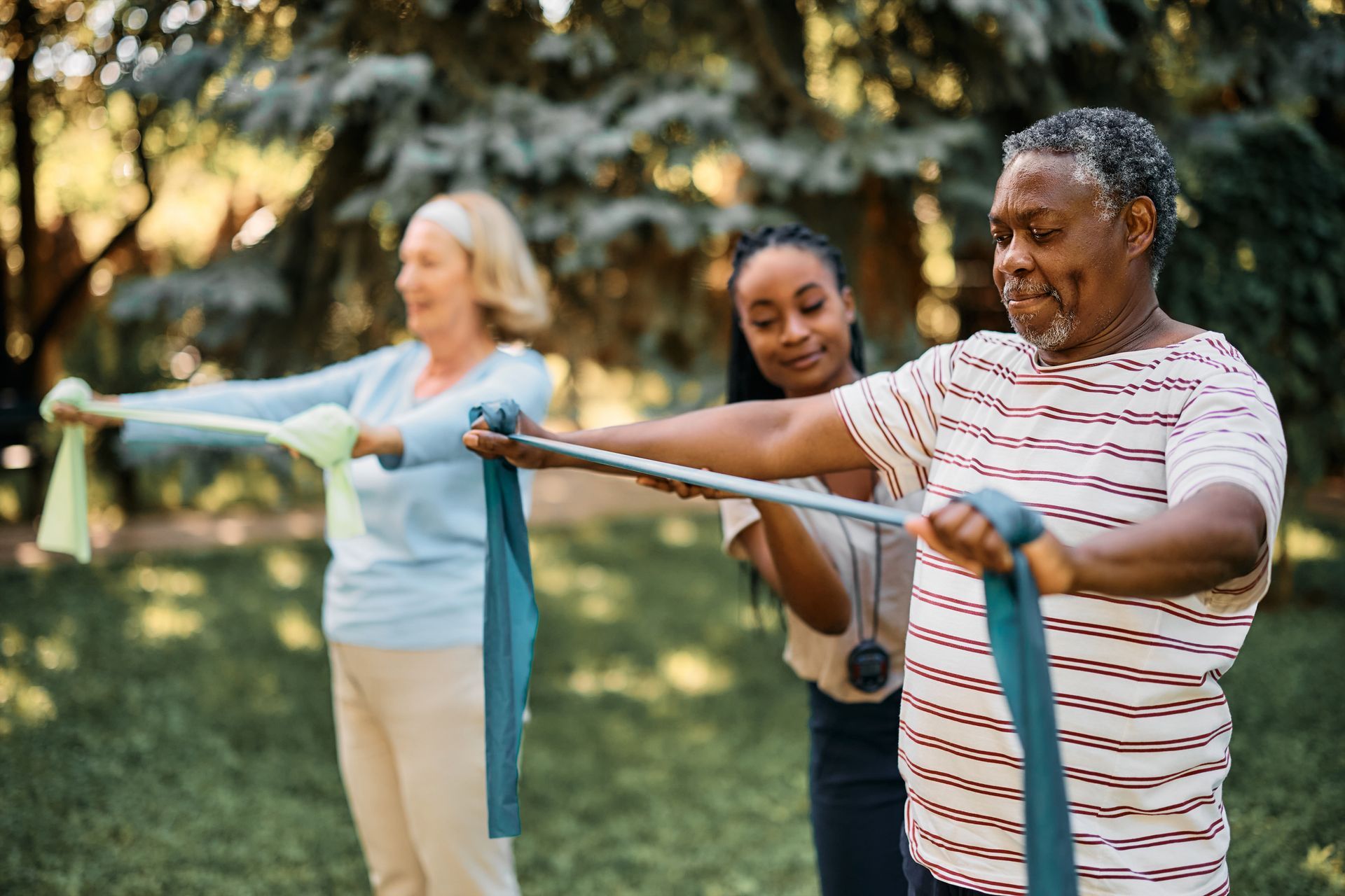 An older Black man and a white woman doing exercises with resistance bands, supervised by a young Black woman in a park.